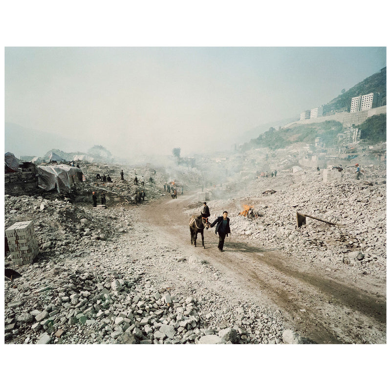 Shot in 2002, Canadian photographer Edward Burtynsky, captures "Feng Jie #5". Depicting a demolished landscape, the photograph is marked by piles of rubble and a dusty haze that obscures the background. Centered in the middle of the work is a man leading his donkey down a dirt road amidst the destruction.