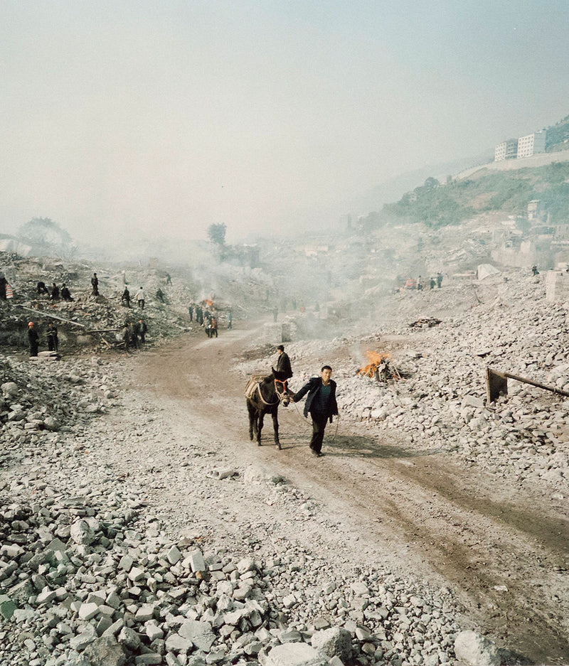 Shot in 2002, Canadian photographer Edward Burtynsky, captures "Feng Jie #5". Depicting a demolished landscape, the photograph is marked by piles of rubble and a dusty haze that obscures the background. Centered in the middle of the work is a man leading his donkey down a dirt road amidst the destruction.