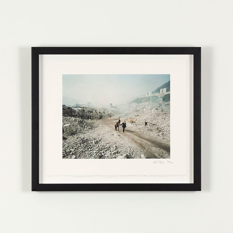 Shot in 2002, Canadian photographer Edward Burtynsky, captures "Feng Jie #5". Depicting a demolished landscape, the photograph is marked by piles of rubble and a dusty haze that obscures the background. Centered in the middle of the work is a man leading his donkey down a dirt road amidst the destruction.