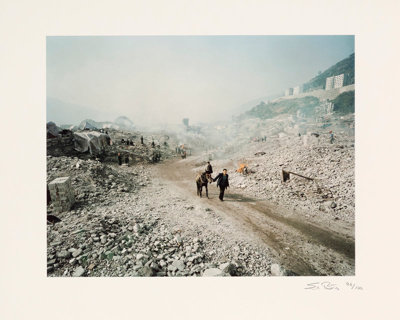 Shot in 2002, Canadian photographer Edward Burtynsky, captures "Feng Jie #5". Depicting a demolished landscape, the photograph is marked by piles of rubble and a dusty haze that obscures the background. Centered in the middle of the work is a man leading his donkey down a dirt road amidst the destruction.